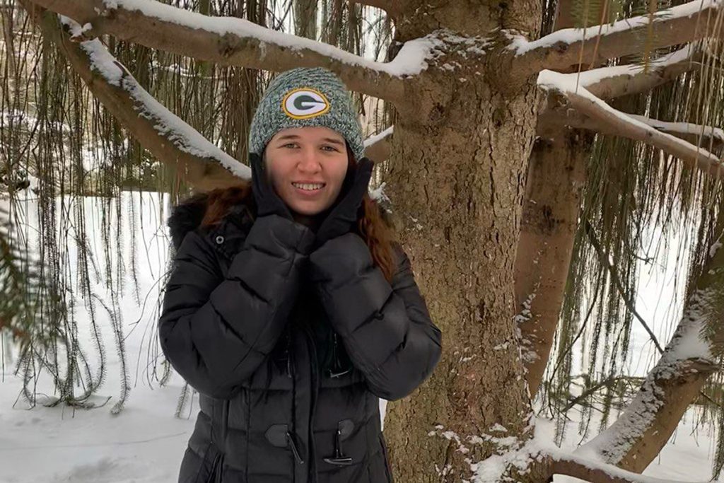 A white woman standing in front of a bare tree. The tree and surrounding area have snow on them as it is the middle of winter. The woman is wearing a black coat while wearing a Green Bay Packers beanie. She has her hands on her cheeks.