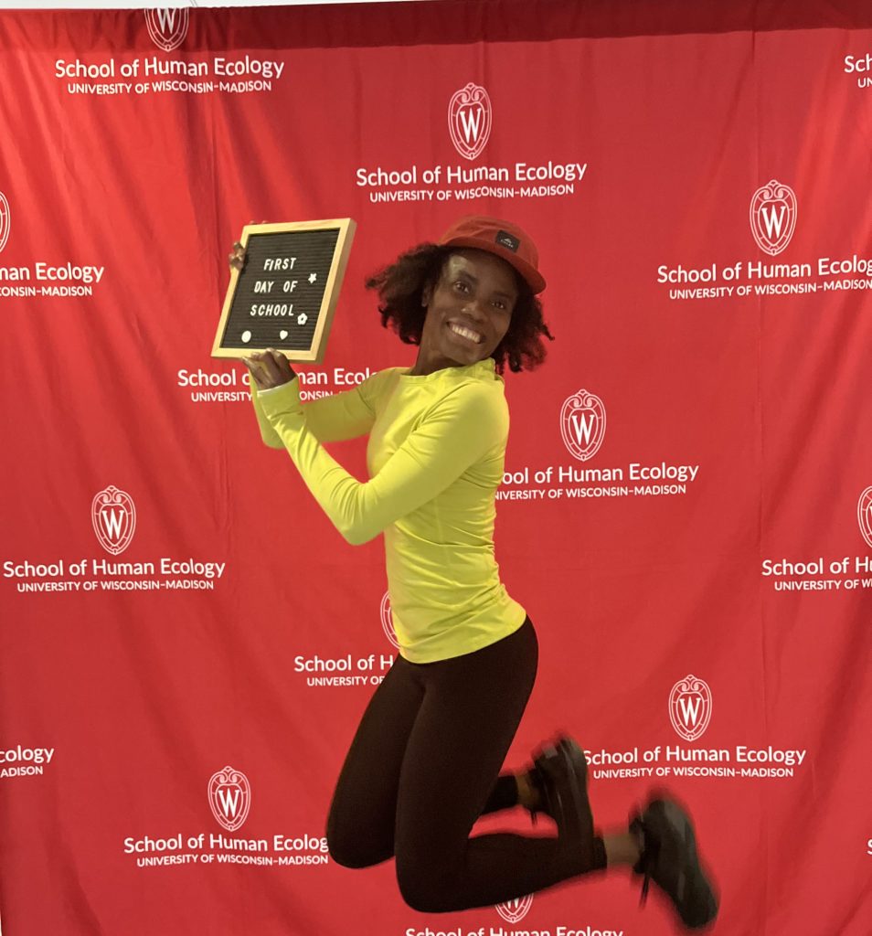 A black woman wearing a bright yellow long-sleeve shirt, black pants, and black shoes is jumping in the air holding a sign that says "First Day of School"