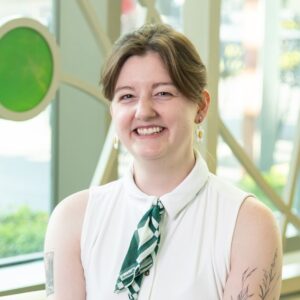 A white woman with short brown hair smiling for a headshot portrait photo. She is wearing daisy flower earrings and a white sleeveless shirt with a green ascot in the collar.