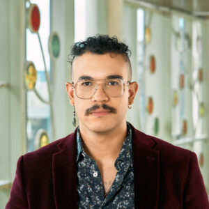 A mixed-race, brown, nonbinary person, with a curly dark brown mohawk and mustache, wearing a red velvet blazer and dark flowered shirt.