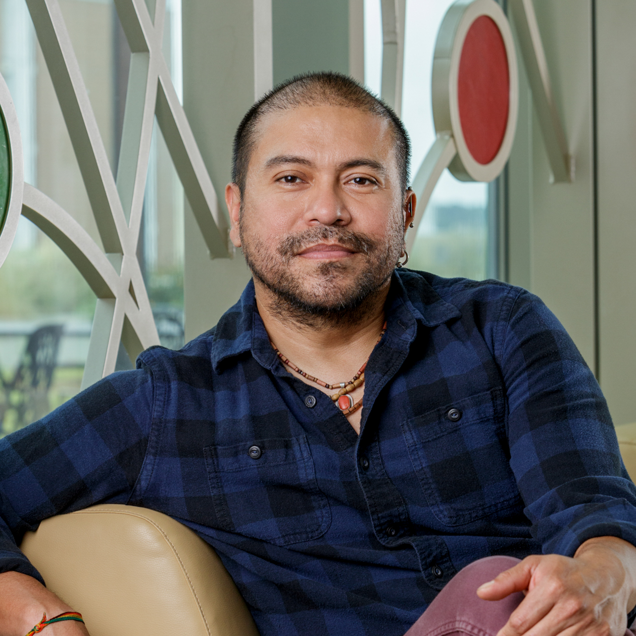 A latino man with a buzz cut poses for a headshot portrait photo. He is wearing a dark blue and black, checkered, flannel shirt. He also has some necklaces and bracelets on. He has a short beard.