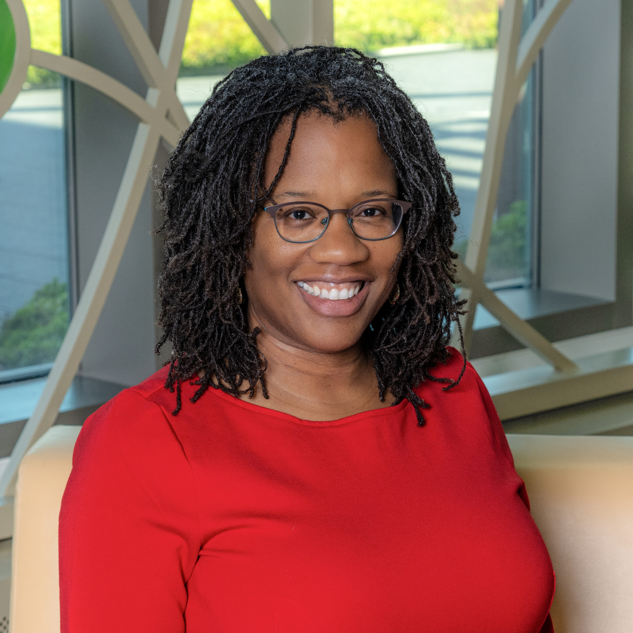 African American woman with shoulder length hair wearing a red long-sleeve shirt and black glasses, smiles for a portrait shot
