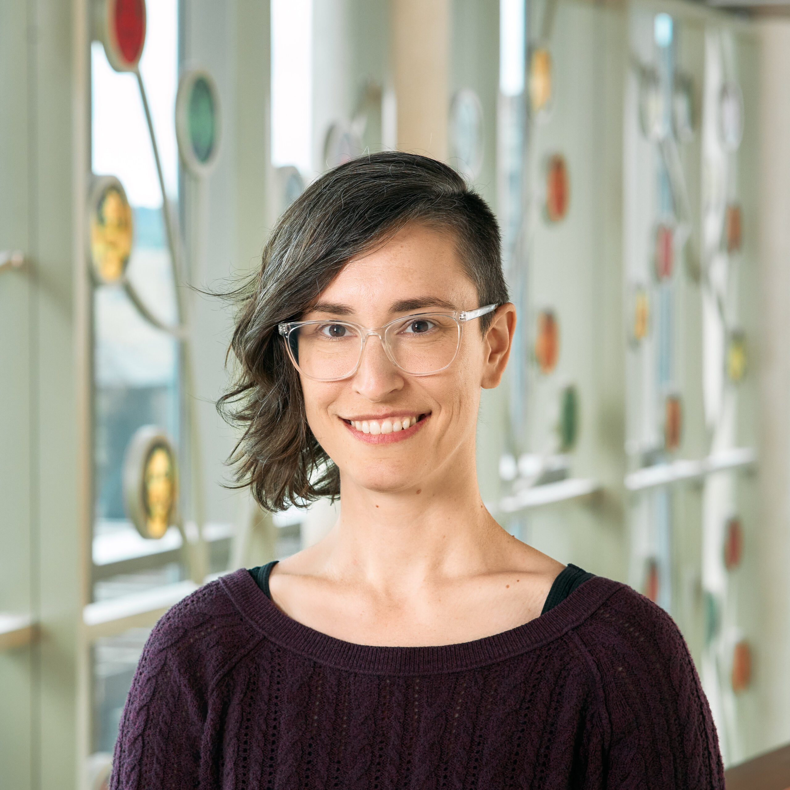 A white woman smiling with brown eyes and wavy brown hair that is shaved on one side, wearing clear plastic frame glasses and a purple sweater.