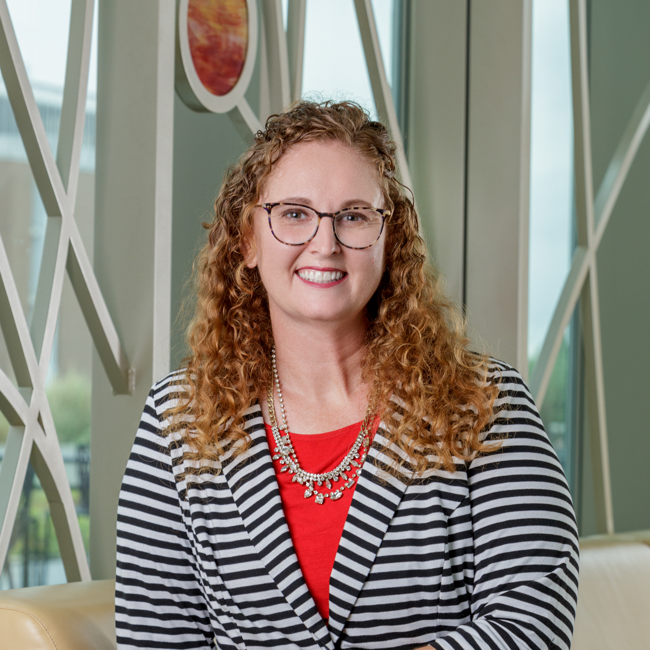 White woman smiling for a headshot portrait. She has curly, red hair that falls below her shoulders. She is wearing black and brown frame glasses and a black and white striped blazer with a red shirt underneath. She is also wearing two necklaces around her neck. 