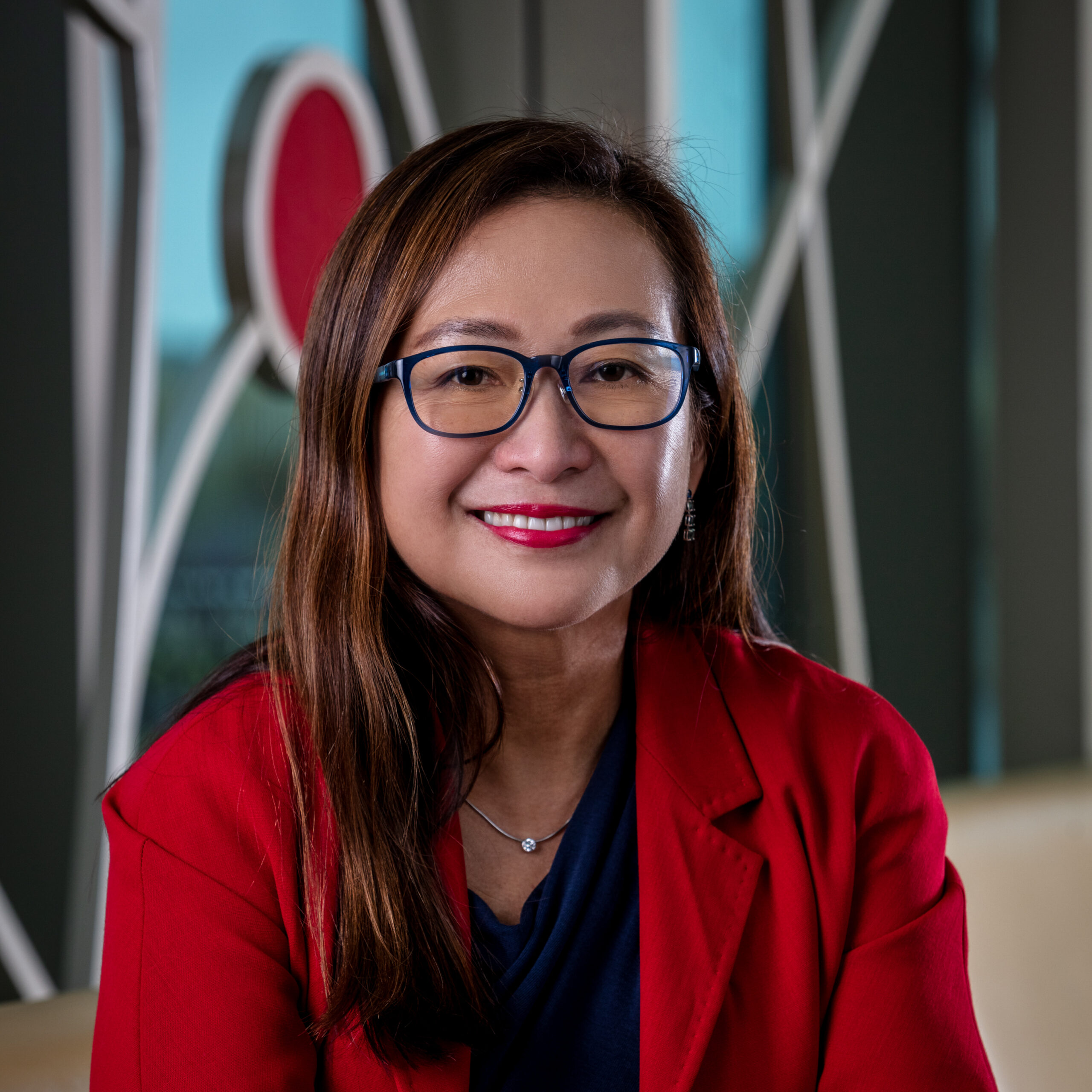 Asian woman with long brown hair, wearing a red blazer and dark blue glasses, smiling for a portrait shot